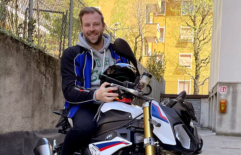 Man sitting on a motorbike in a sunny courtyard holding a helmet