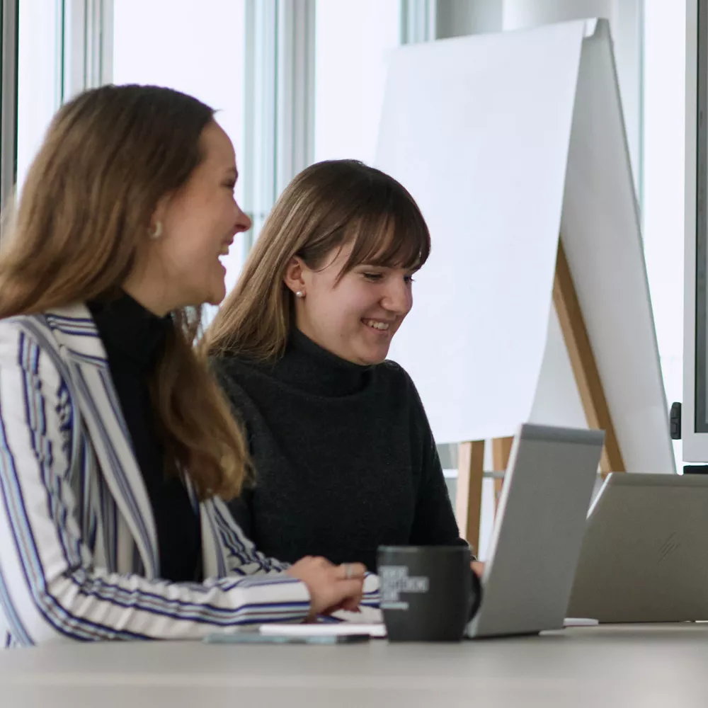 Two female colleagues sit next to each other at their laptops and work smilingly